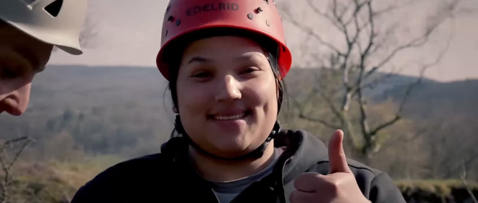 Een jonge deelnemer met rode helm steekt lachend een duim op, met de vallei op de achtergrond.